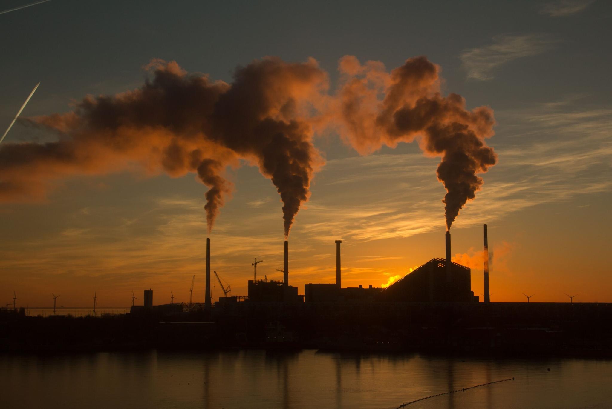 Plano de Descarbonização Industrial Chaminés industriais emitindo fumaça durante o pôr do sol, com turbinas eólicas ao redor da planta.