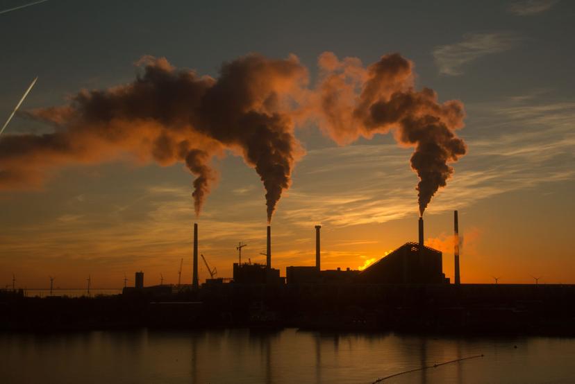 Plano de Descarbonização Industrial Chaminés industriais emitindo fumaça durante o pôr do sol, com turbinas eólicas ao redor da planta.