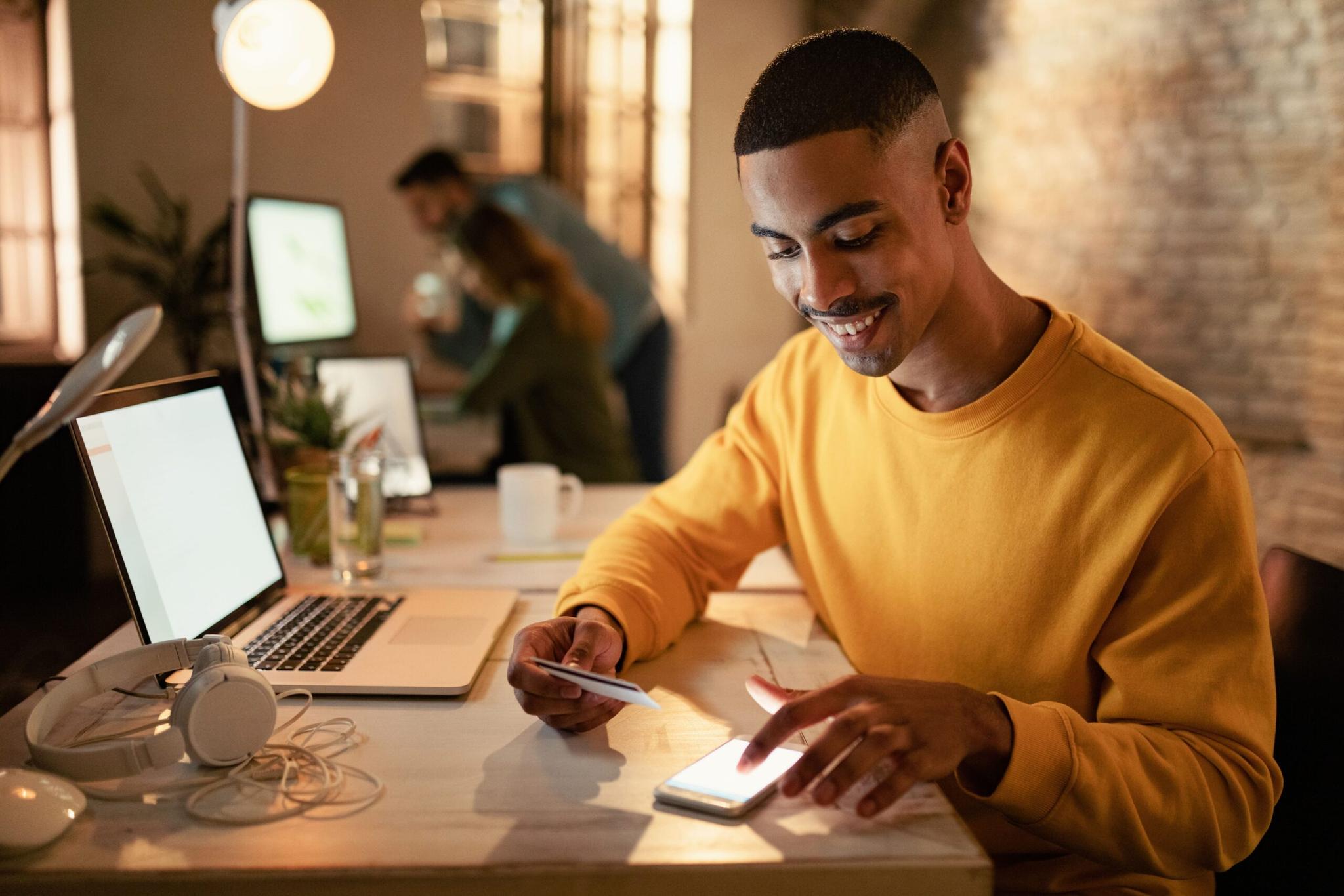 fomento-de-inovação-para-fintechs Homem de blusa amarela sorri ao digitar no celular e segurar cartão de crédito em mesa com laptop em escritório compartilhado.