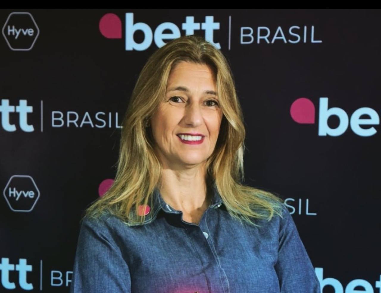 Mulher com longos cabelos loiros, vestindo uma camisa jeans e sorrindo, tendo como fundo a Bett Brasil.