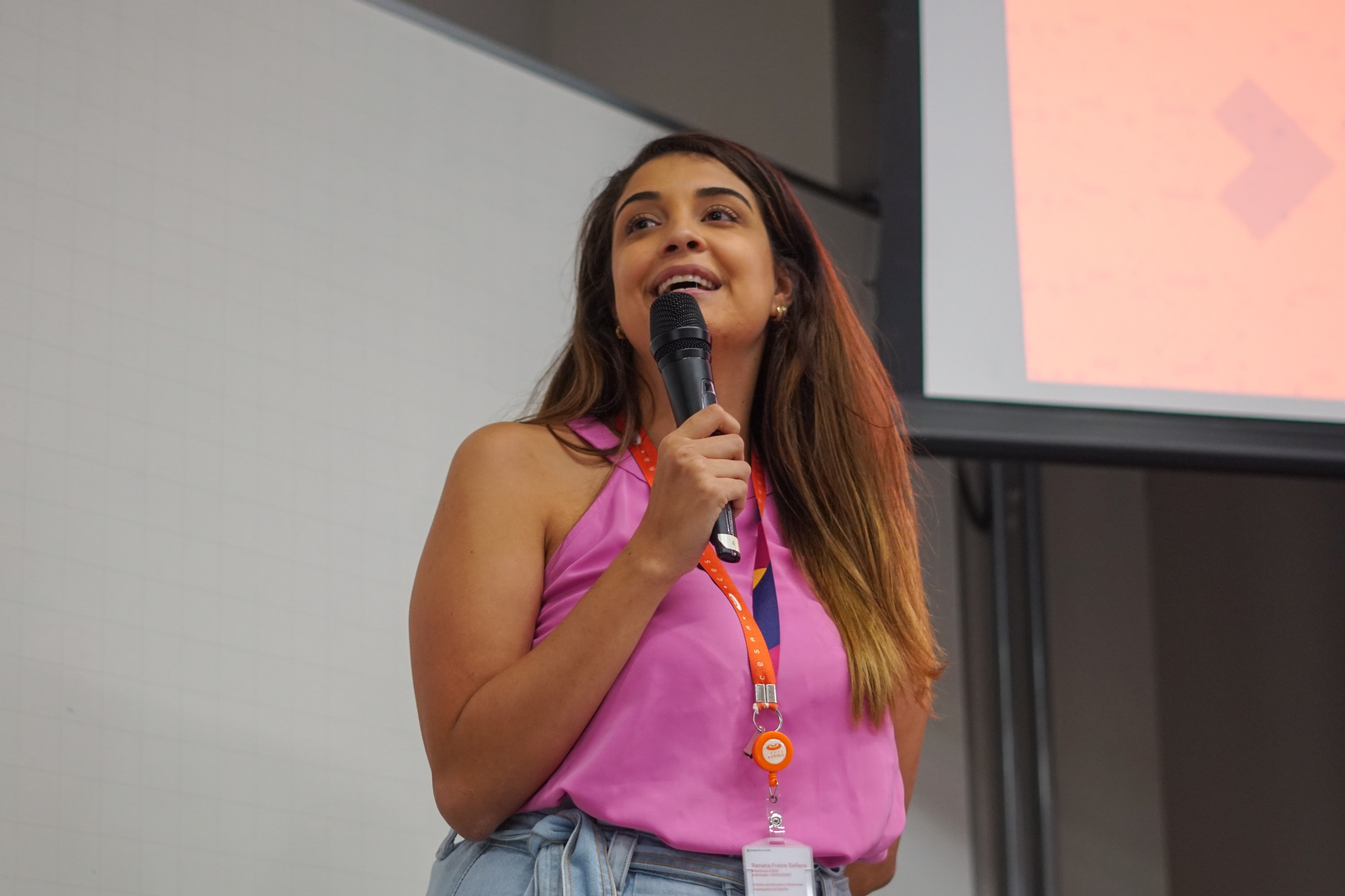 Mulher falando ao microfone, vestindo uma blusa rosa e sorrindo, em pé na frente de uma tela de apresentação.