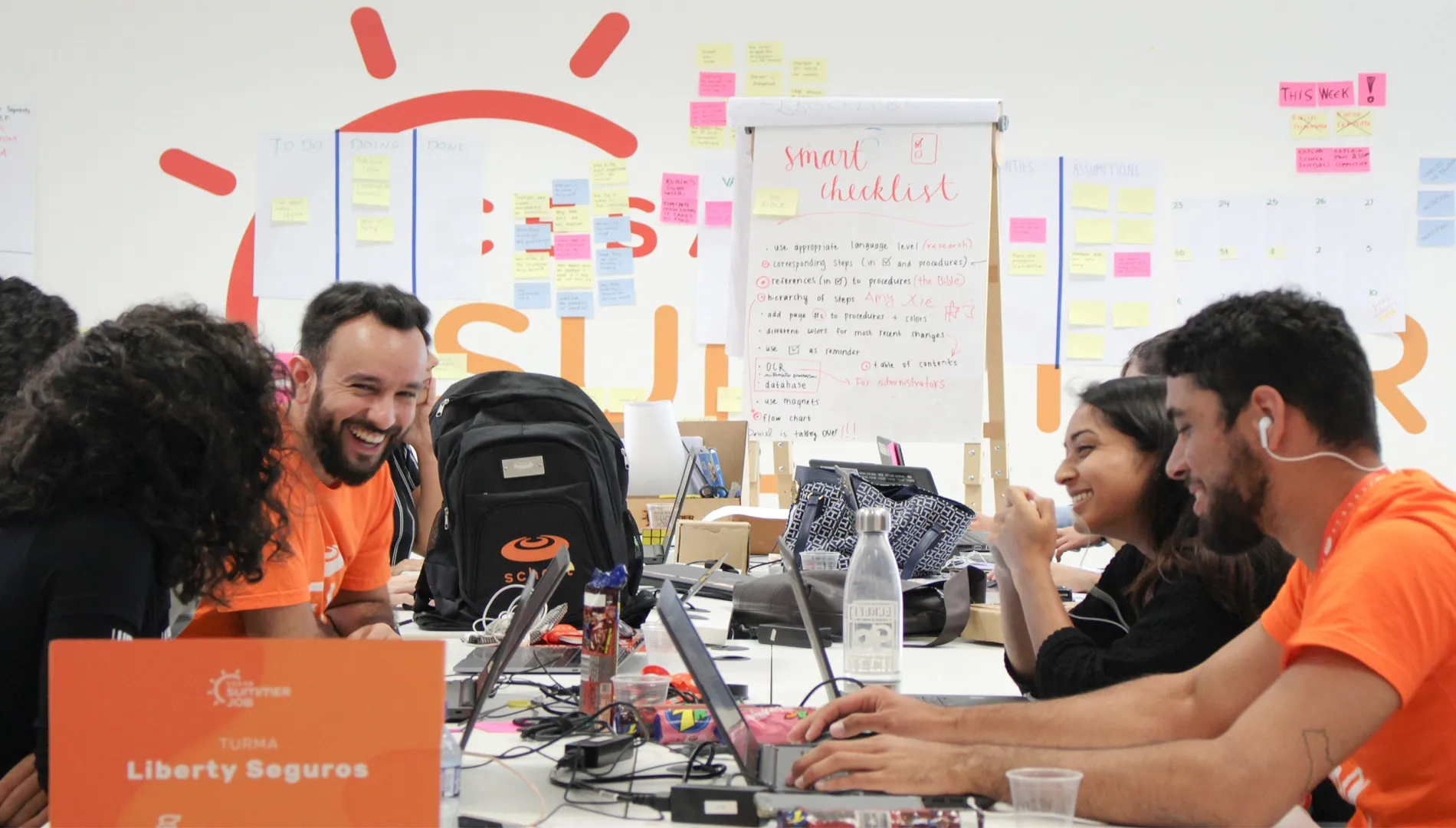 Jovens em volta de mesa com notebooks e post-its ao fundo. Também há uma placa que identifica turma Liberty Seguros no Summer Job
