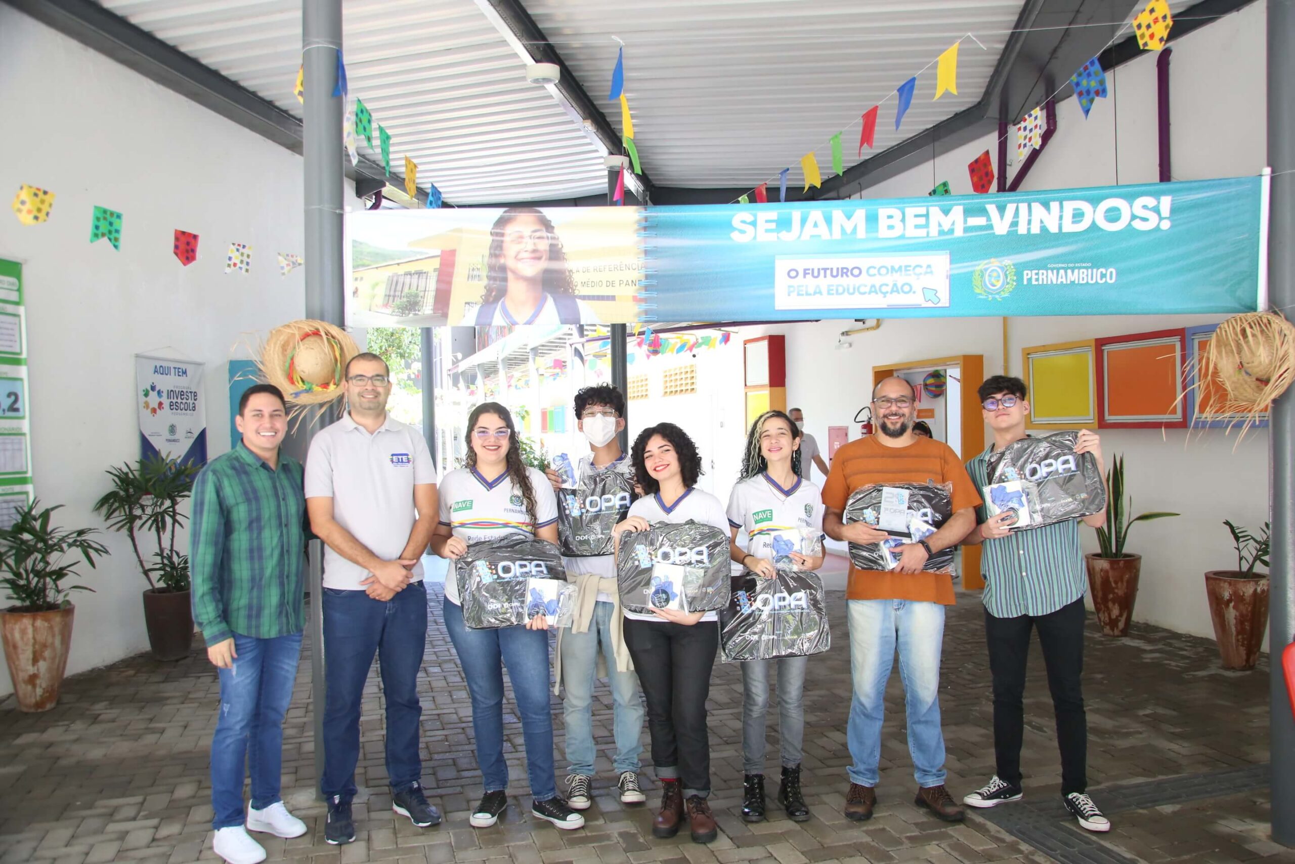 Grupo de estudantes e professores em escola decorada para festa, recebendo mochilas e kits escolares. Banner de boas-vindas e decoração de bandeirinhas.