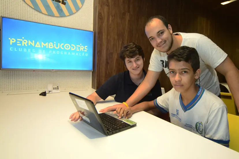 Três jovens sorriem ao redor de um portátil numa mesa branca. Atrás, um ecrã exibe o logótipo amarelo Pernambucoders