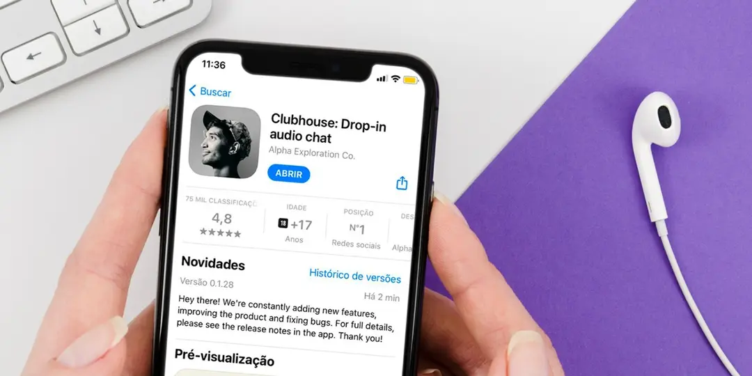 Mãos seguram celular exibindo a página do app Clubhouse na App Store, ao lado de um fone de ouvido branco