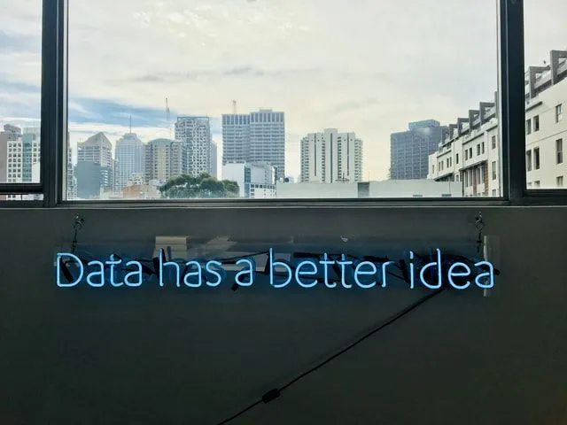 Letreiro de neon azul com a frase "Data has a better idea" fixado abaixo de uma janela com vista para prédios