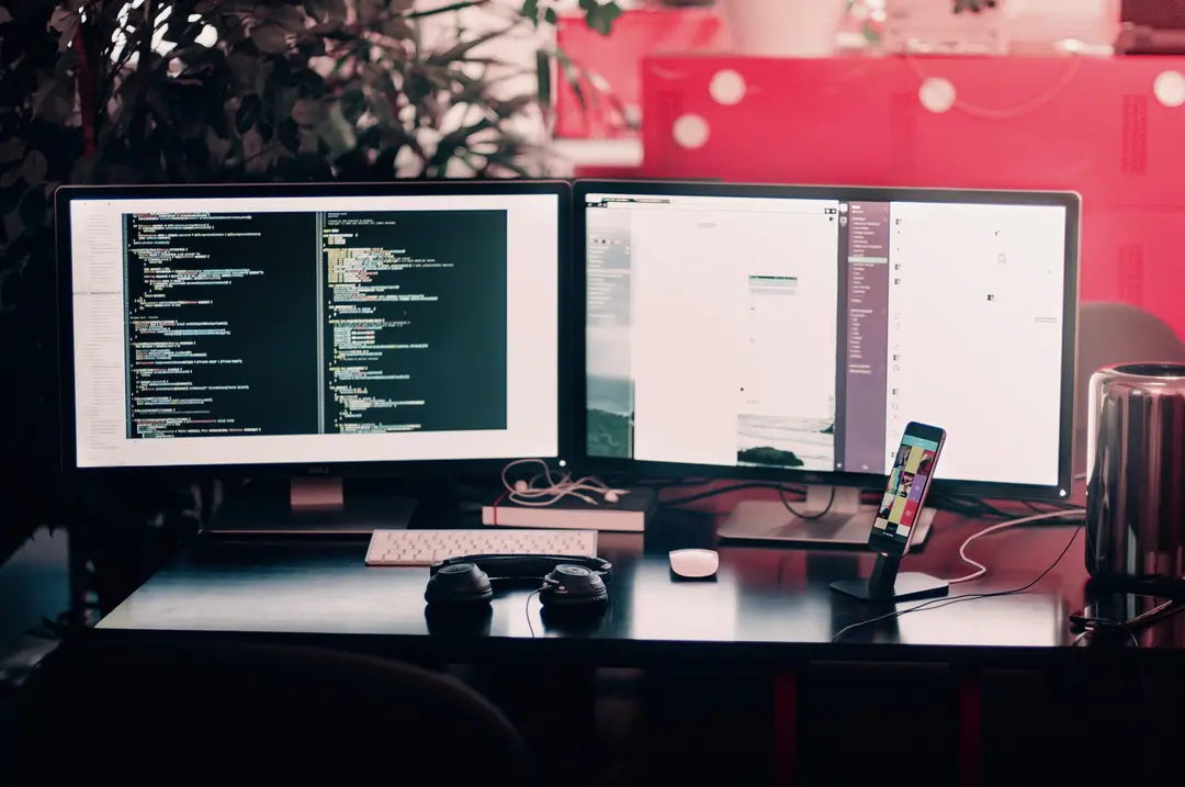 Dois monitores acesos mostram códigos e sistemas em uma mesa com teclado, mouse, fone de ouvido e celular