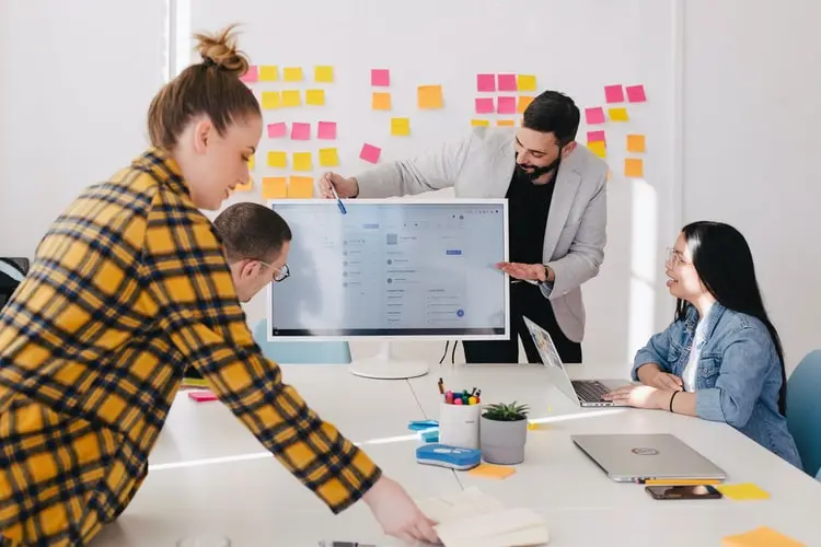 Quatro pessoas debatem ideias ao redor de uma mesa com notebooks e uma grande tela, diante de um painel de post-its