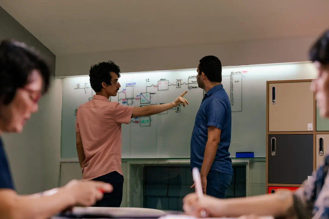 Dois profissionais debatem em pé diante de uma grande lousa de vidro com diagramas e fluxogramas desenhados, enquanto outros colegas aparecem em primeiro plano