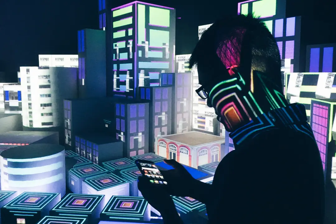 Homem de óculos usa smartphone de perfil diante de maquete de cidade futurista iluminada por luzes neon coloridas.