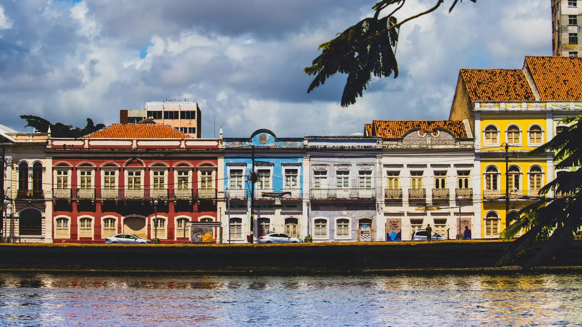 Casarões históricos coloridos na beira do rio no Recife Antigo sob céu nublado, vistos por trás de folhagens.