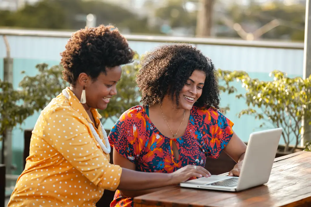 Duas mulheres negras sorridentes sentadas em mesa externa de madeira usam um laptop juntas em um ambiente com plantas.