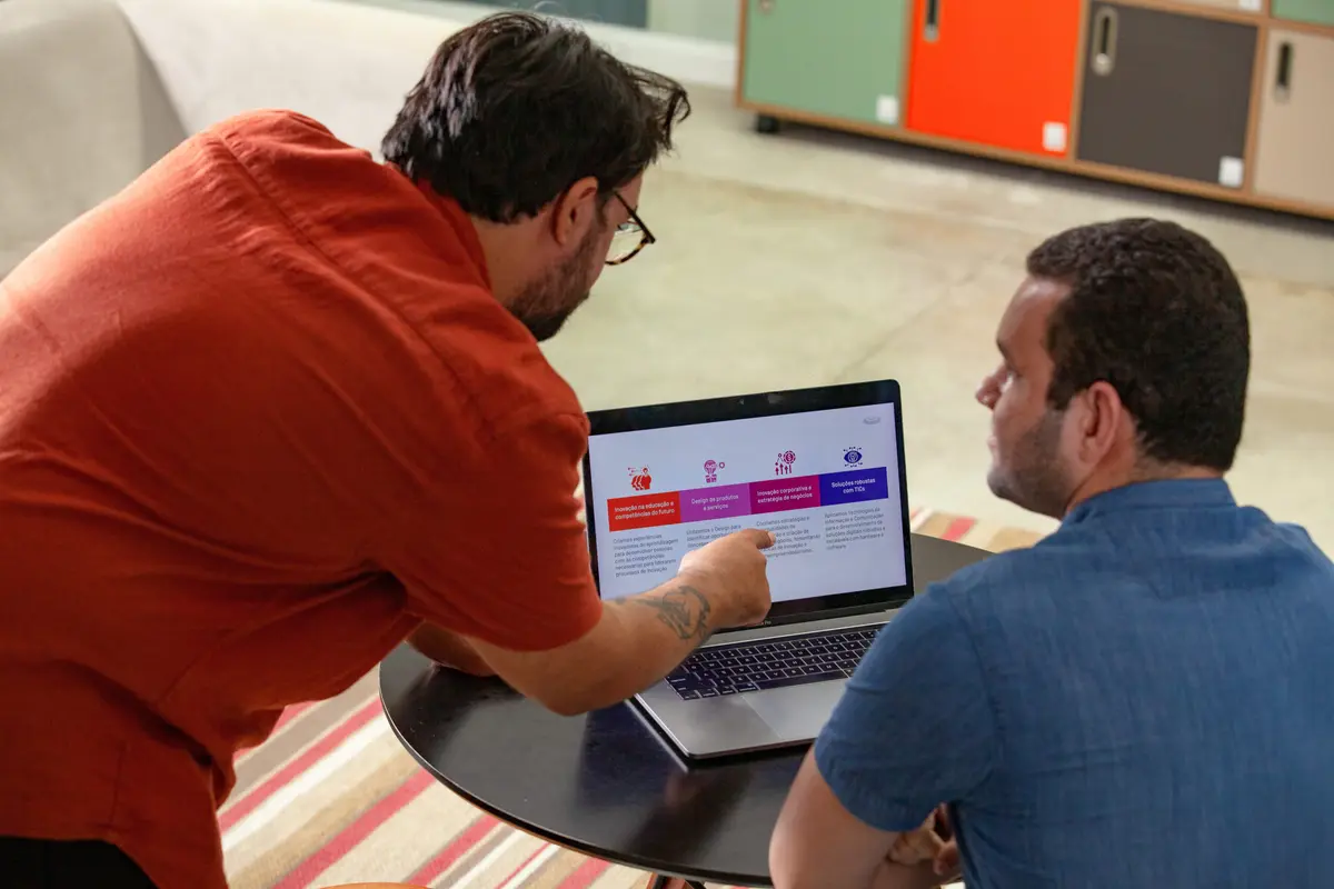 Dois homens analisam notebook com apresentação sobre inovação e estratégia. Um aponta para a tela enquanto o outro observa.