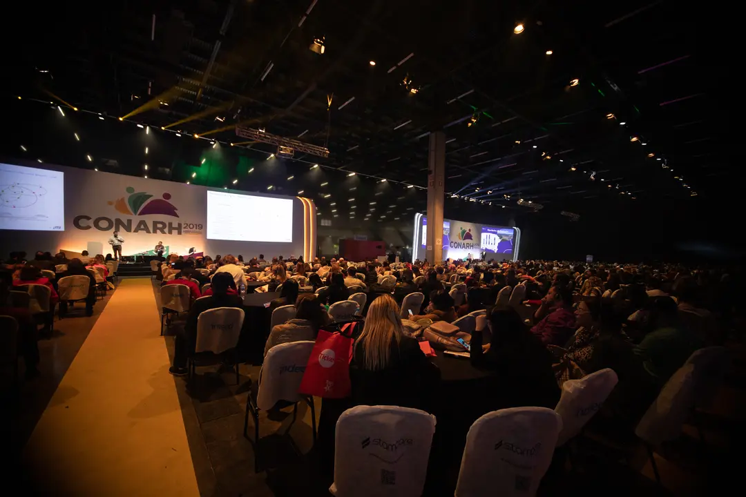 Grande público assistindo a uma palestra no evento CONARH com palestrante no palco e dois grandes telões iluminados ao fundo.