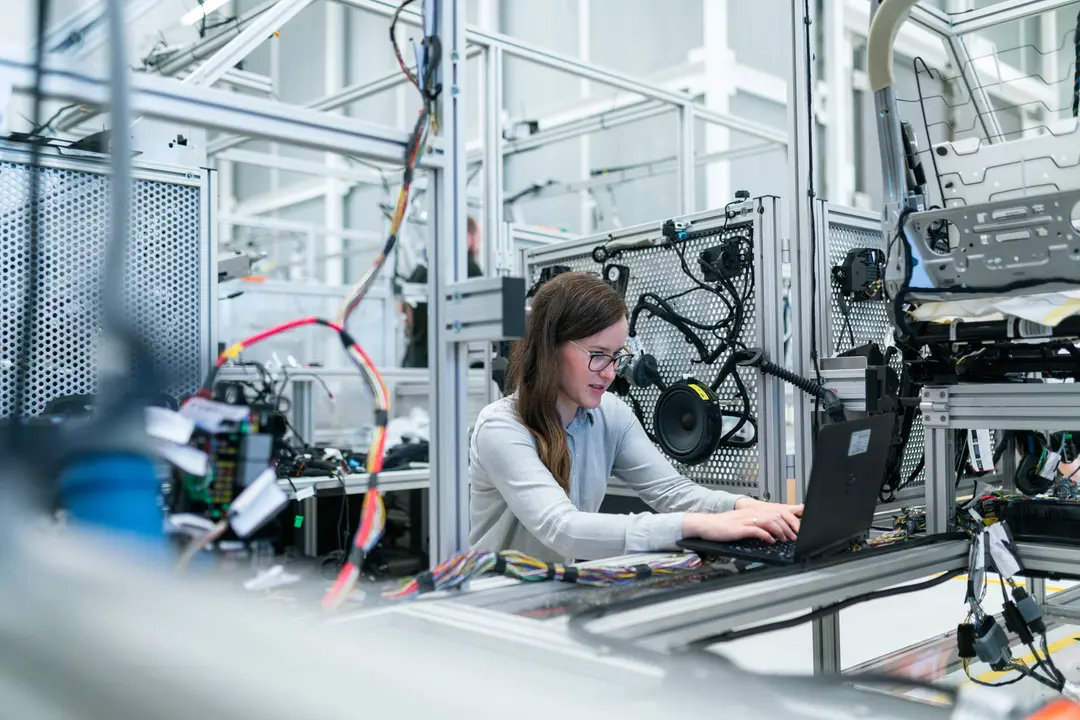 Mulher de óculos operando um laptop em bancada de laboratório industrial com fiações e estruturas metálicas ao redor.