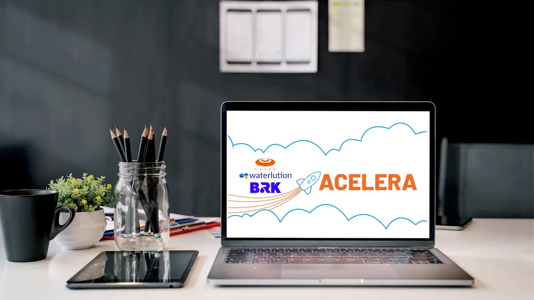 Notebook aberto em mesa de escritório exibe o logo Acelera com ícone de foguete, marcas BRK, Waterlution e CESAR ao fundo.