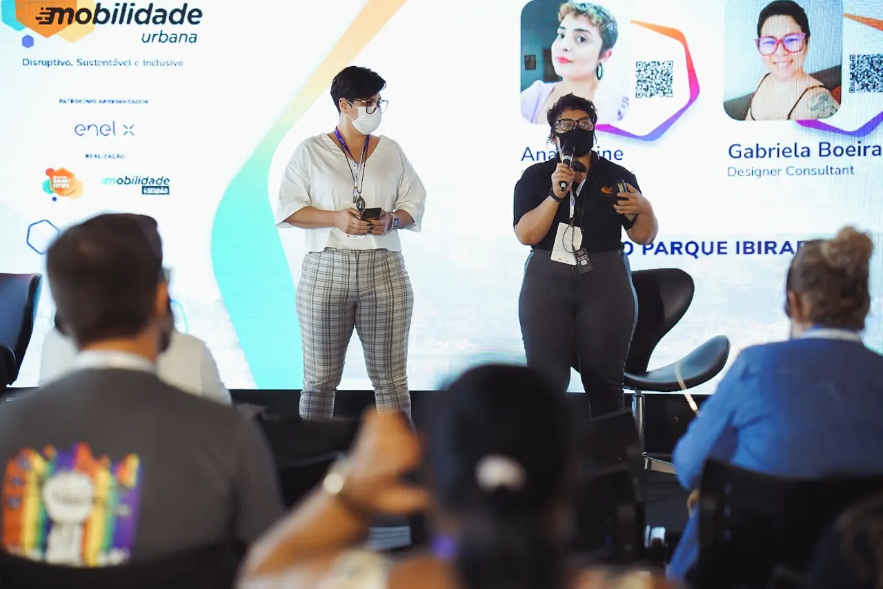 Duas mulheres de máscara palestram em palco diante de telão sobre mobilidade urbana, enquanto o público assiste de costas.