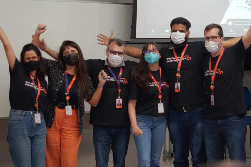 Seis profissionais usando máscaras e camisetas pretas comemoram com braços levantados em frente a uma tela de projeção