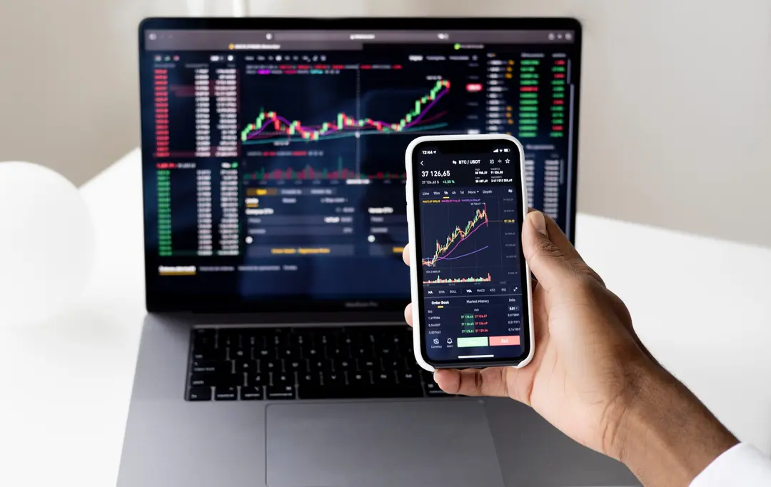 Mão segura um smartphone que exibe um gráfico de velas financeiras do par BTC/USDT em frente à tela de um laptop com gráficos de trade abertos.