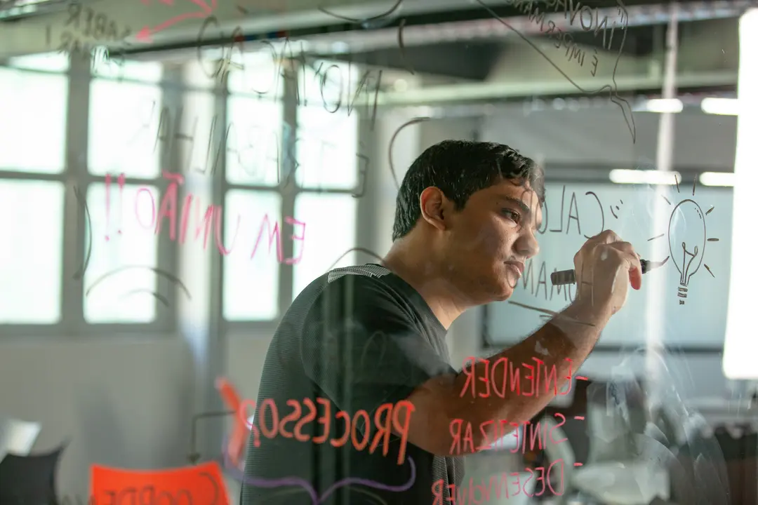 Homem desenha uma lâmpada com piloto preto em uma parede de vidro transparente que possui outras anotações coloridas escritas à mão.