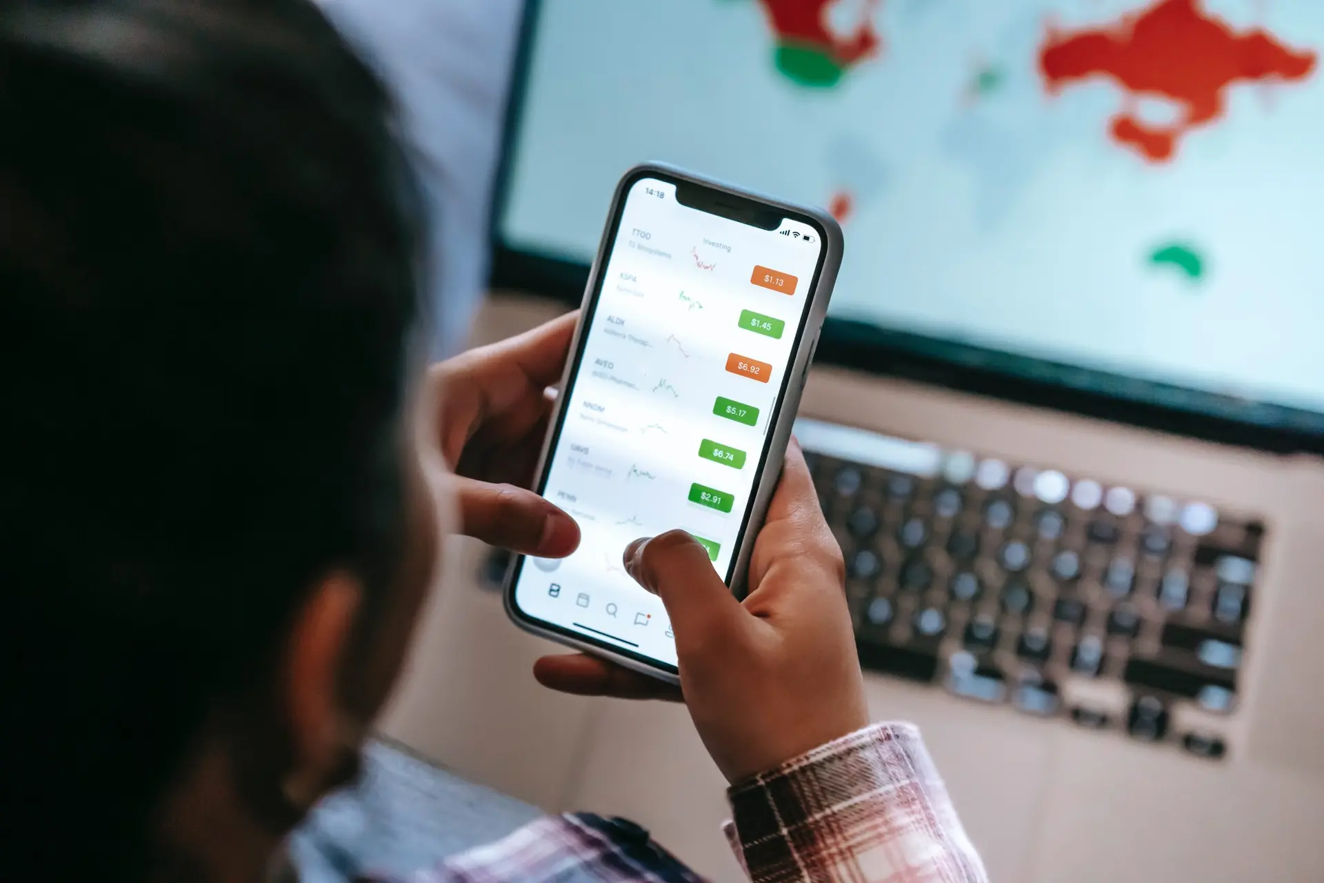 Pessoa segura um celular que exibe gráficos e valores financeiros, com a tela de um notebook ao fundo