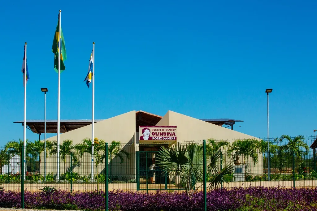 Fachada da Escola Olindina com três bandeiras à esquerda, palmeiras no jardim e céu azul ao fundo sob luz do dia.