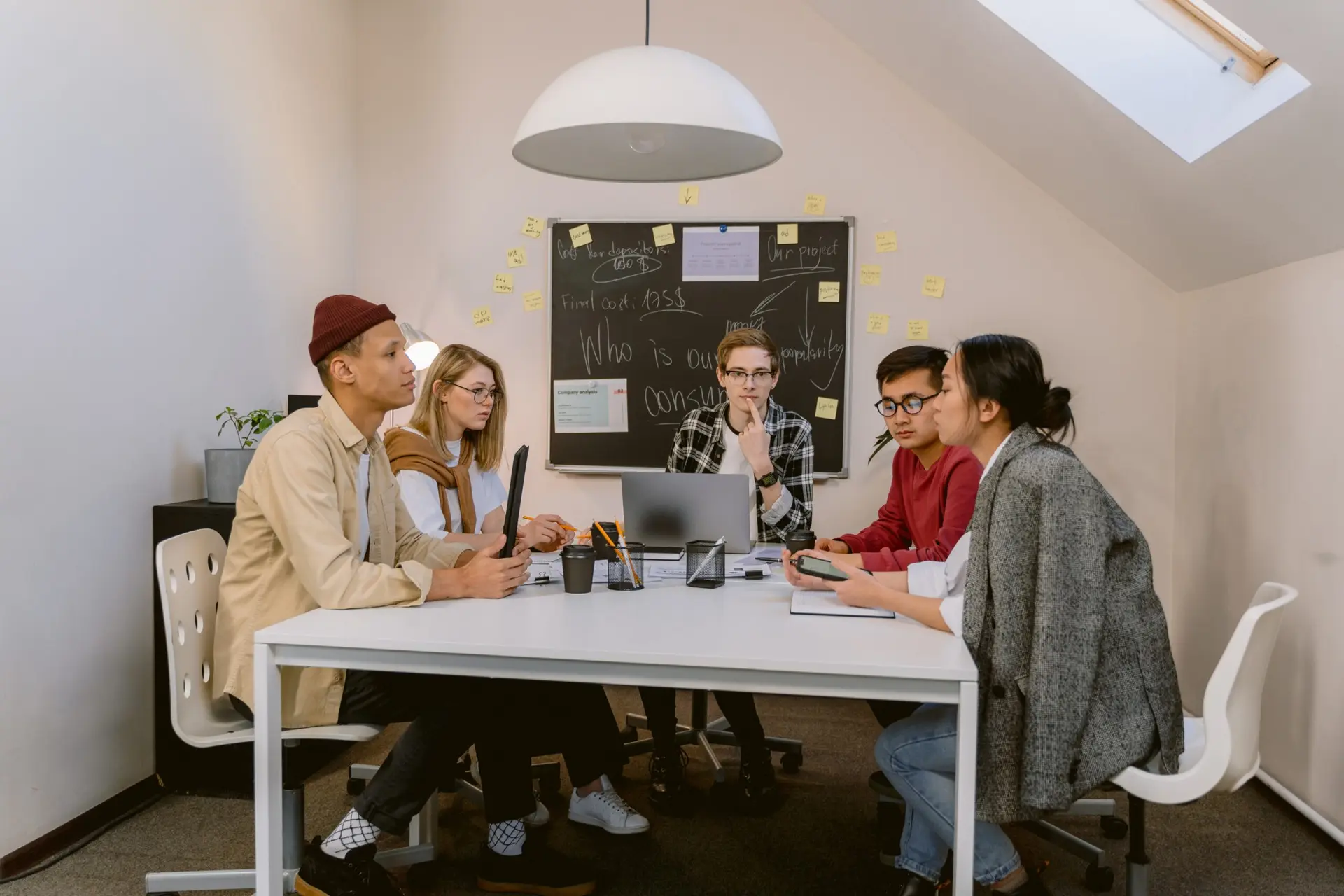 Cinco jovens profissionais estão reunidos ao redor de uma mesa branca de escritório em uma sala de reuniões, conversando e debatendo projetos diante de um quadro-negro com anotações em giz ao fundo