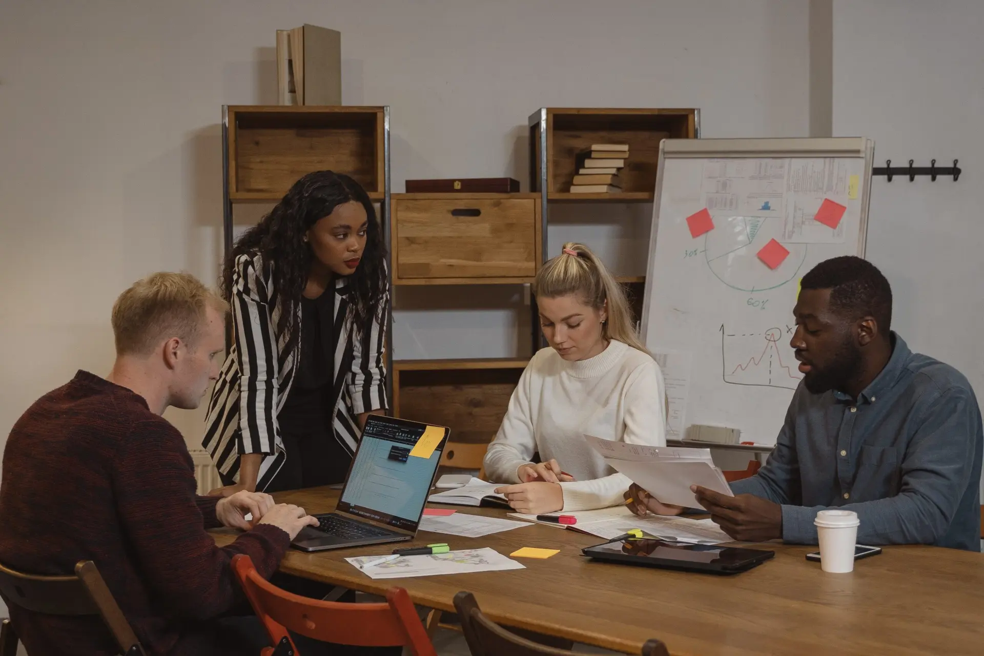 Quatro profissionais em reunião analisando documentos e laptop, com quadro de gráficos e post-its, discutindo inovação e eficiência operacional