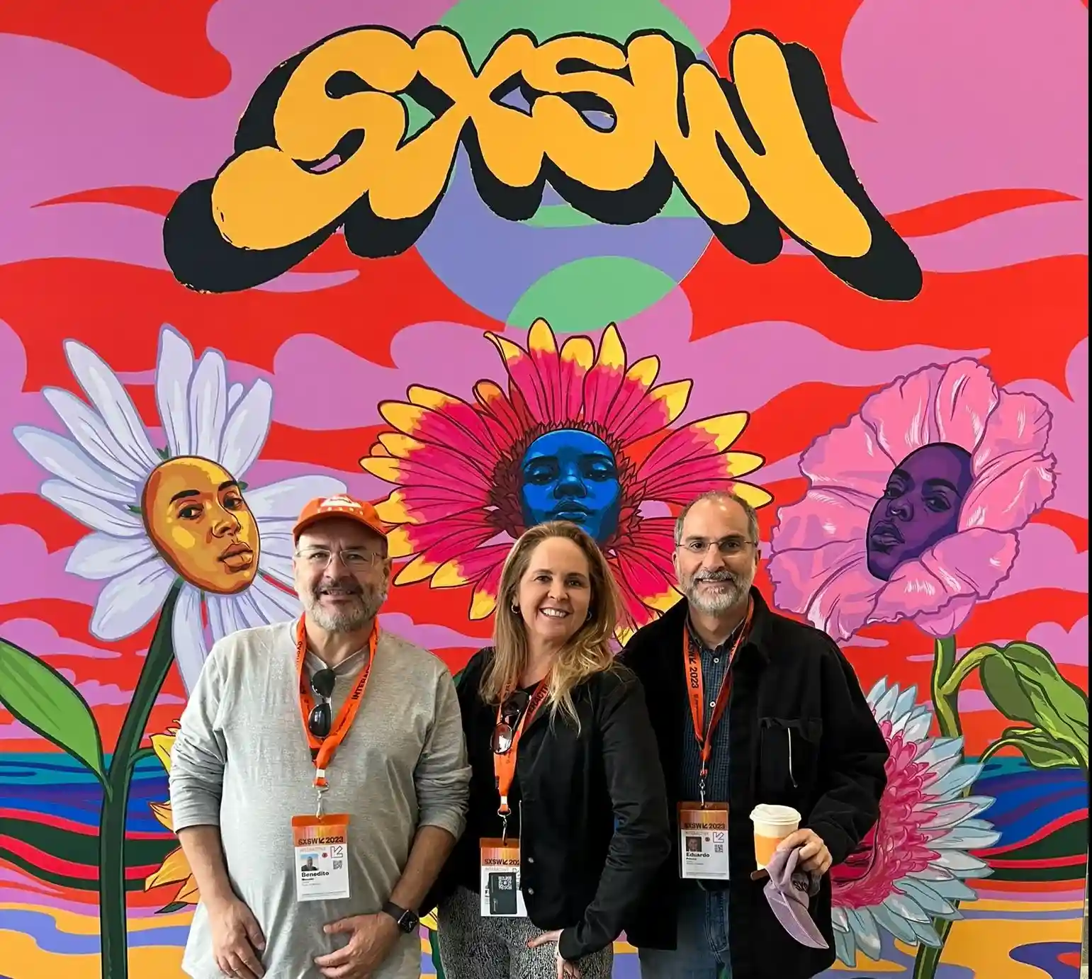 Três pessoas sorrindo em frente a um painel colorido com flores e a logo SXSW, usando crachás do evento em Austin.