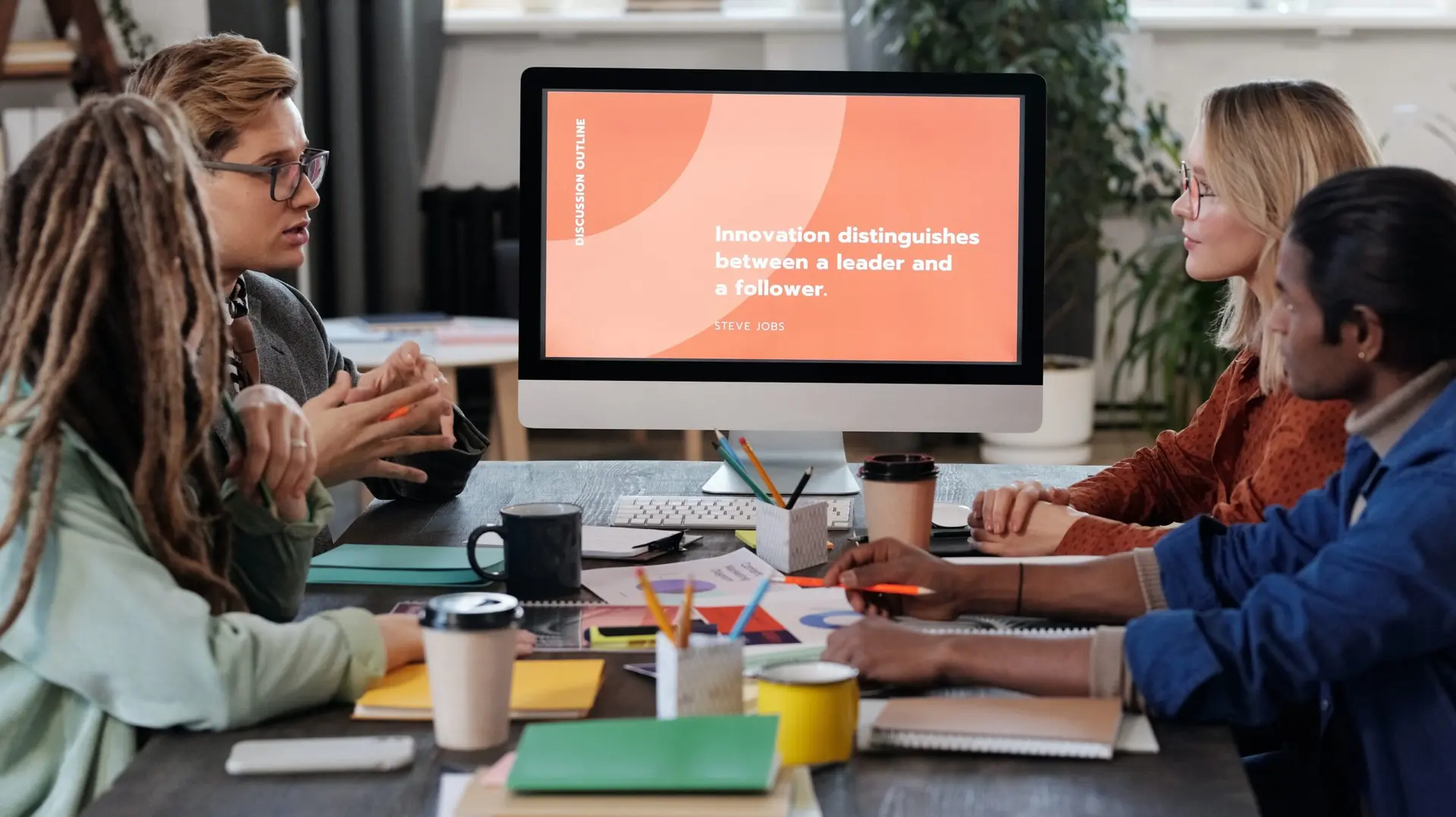 Grupo de quatro pessoas em reunião ao redor de mesa com café, observando monitor com a frase sobre liderança e inovação