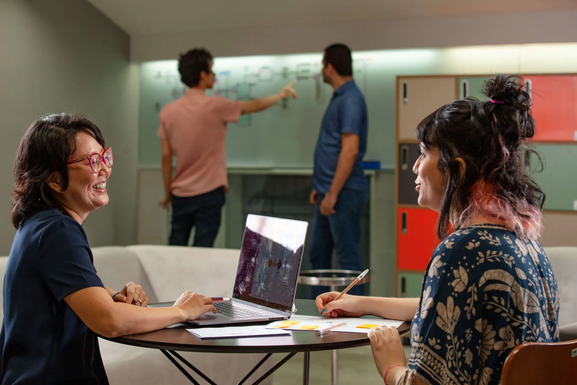 Duas mulheres conversam em uma mesa redonda com laptop e papéis; ao fundo, dois homens discutem diante de um quadro branco.
