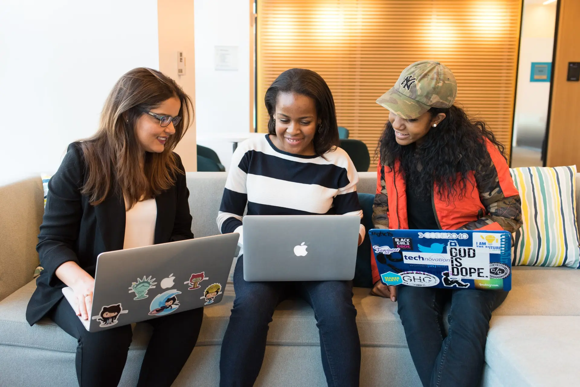Três mulheres sorridentes sentadas em um sofá, cada uma com um laptop, colaborando em um ambiente moderno de escritório.