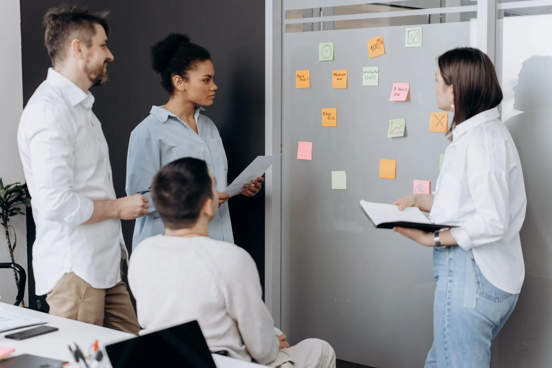 Grupo de quatro profissionais colabora em frente a um quadro de vidro com post-its coloridos em reunião de planeamento.