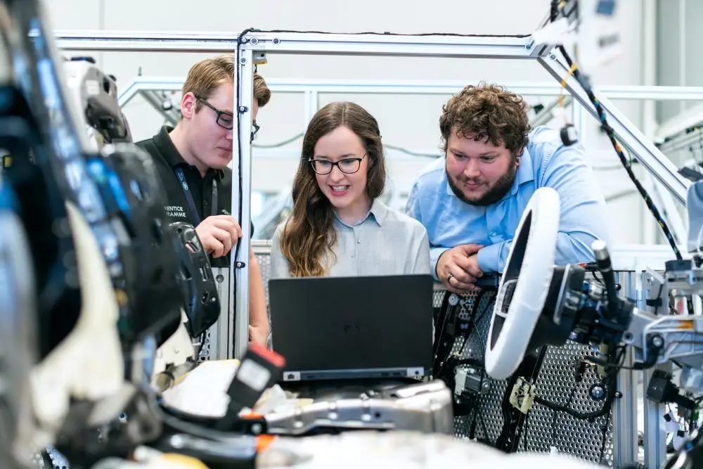 Três engenheiros sorridentes analisam dados em um laptop dentro de um laboratório industrial cercados por cabos e máquinas.