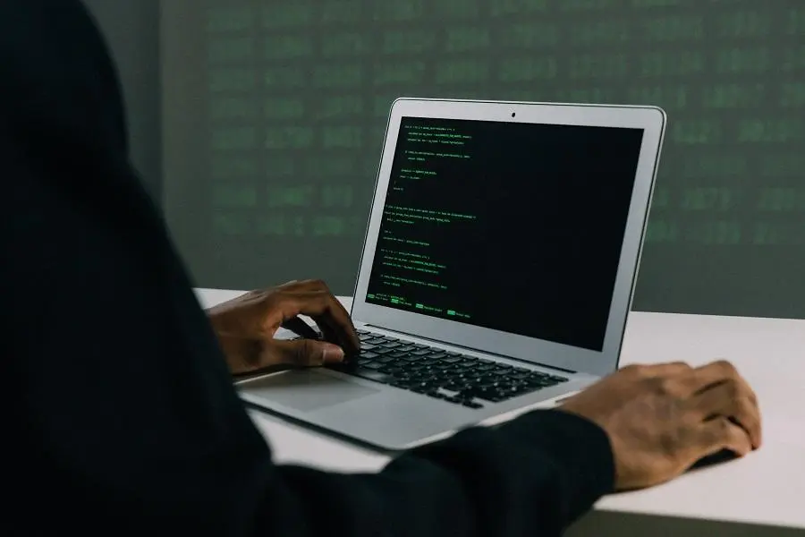 Mãos de uma pessoa programando em um laptop que exibe linhas de código verdes em uma tela preta, em ambiente escuro.