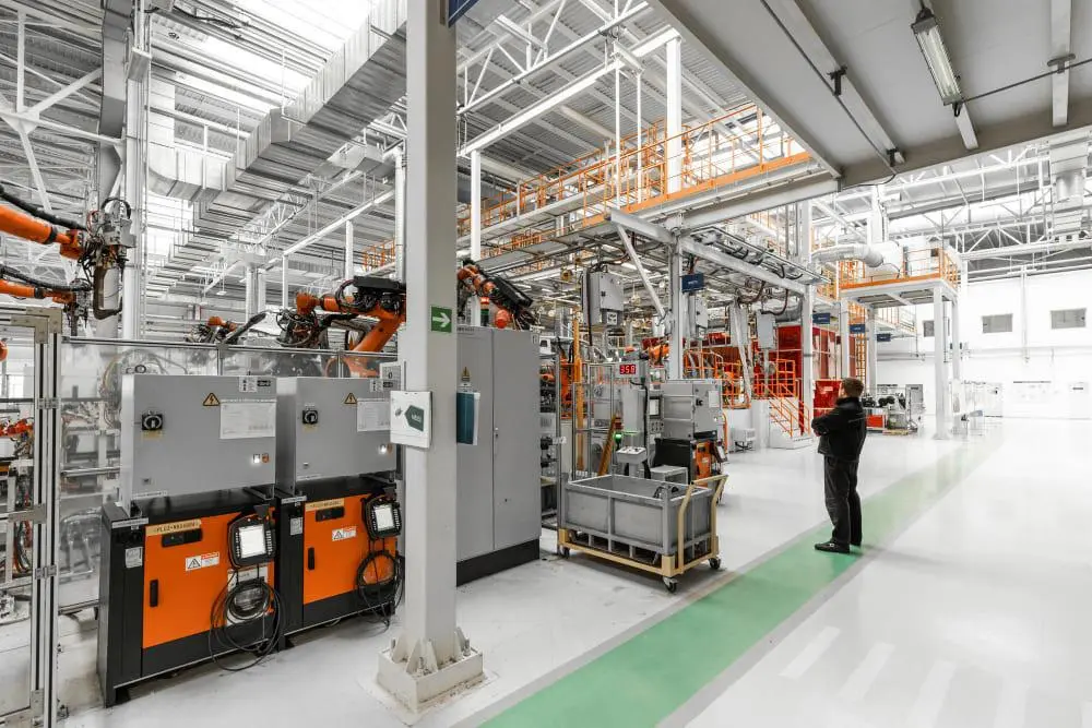 Operário observa braços robóticos em uma ampla fábrica automatizada com painéis de controle laranja e estruturas metálicas.