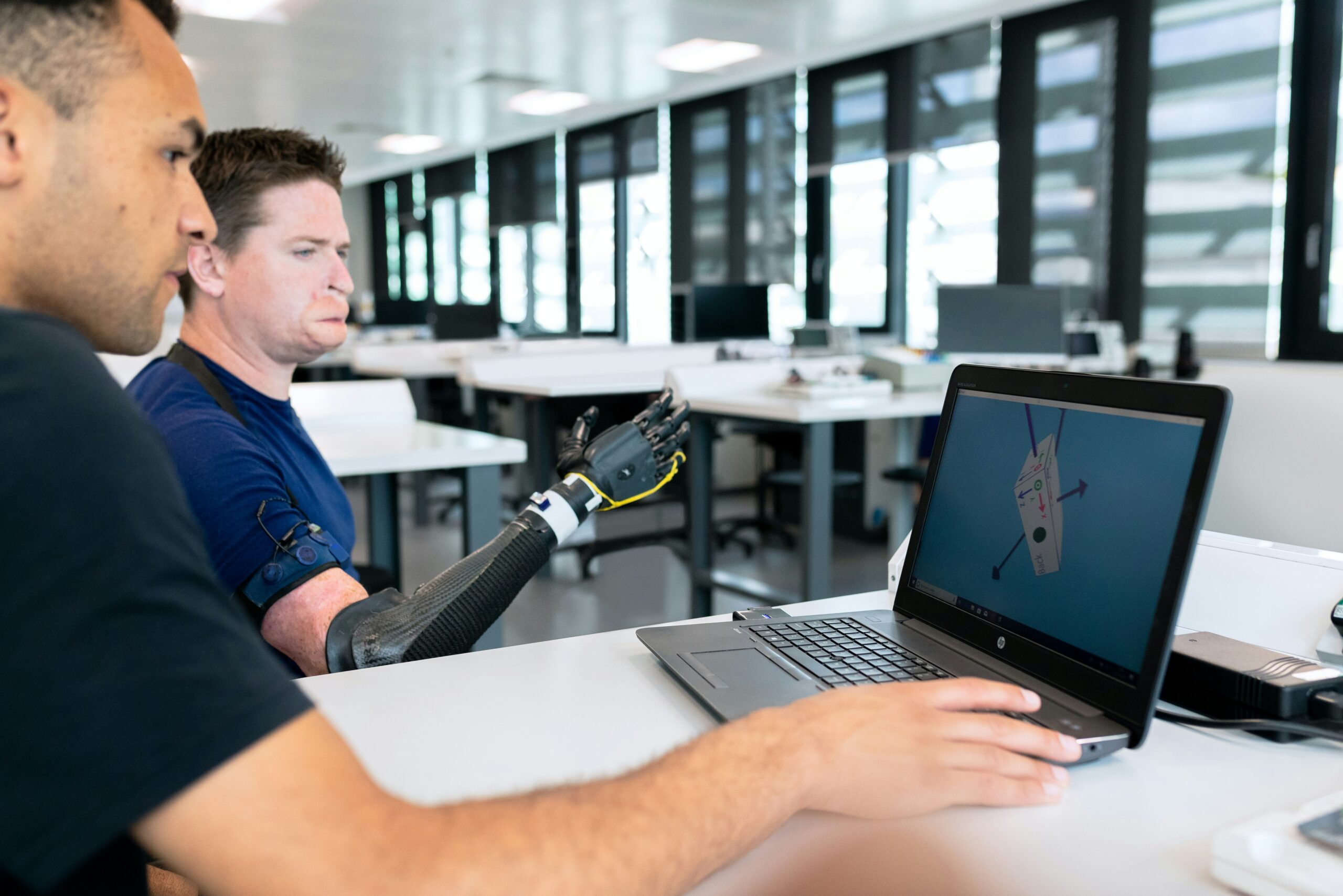 Dois homens sentados a uma mesa, um com uma prótese de braço, usam um laptop que mostra um modelo 3D em um programa de design.