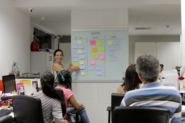 Uma mulher apresenta um quadro branco com post-its coloridos a um grupo de colegas de trabalho sentados em um escritório.