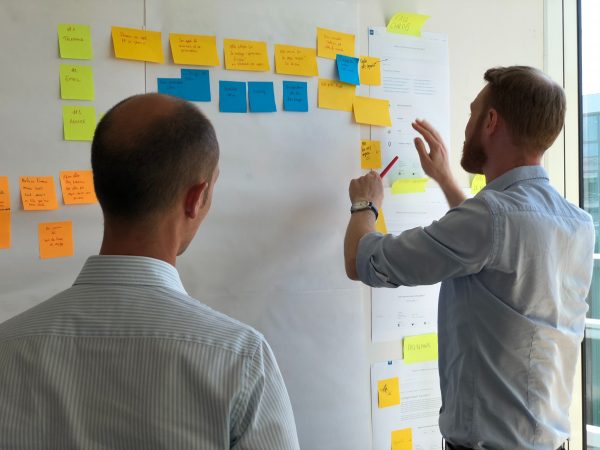 Dois homens em uma sessão de brainstorming, um apontando para um quadro branco coberto de notas adesivas coloridas e documentos.