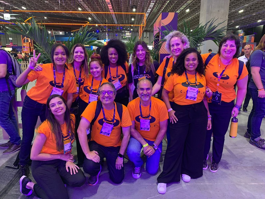 Grupo sorridente vestindo camisetas laranja em evento. Mulheres e homens, em pose descontraída. Algumas pessoas estão agachadas, outras em pé. Ambiente de conferência ou feira.
