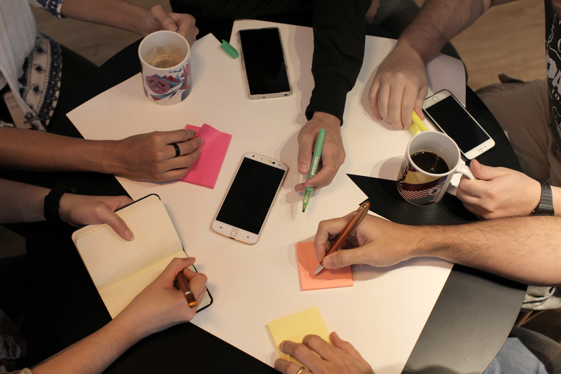 Uma mesa redonda com pessoas sentadas ao redor, compartilhando ideias e trabalhando em um projeto. Smartphones, canetas, blocos de notas e xícaras de café estão espalhados pela mesa branca. Notas adesivas coloridas são usadas para brainstorming.