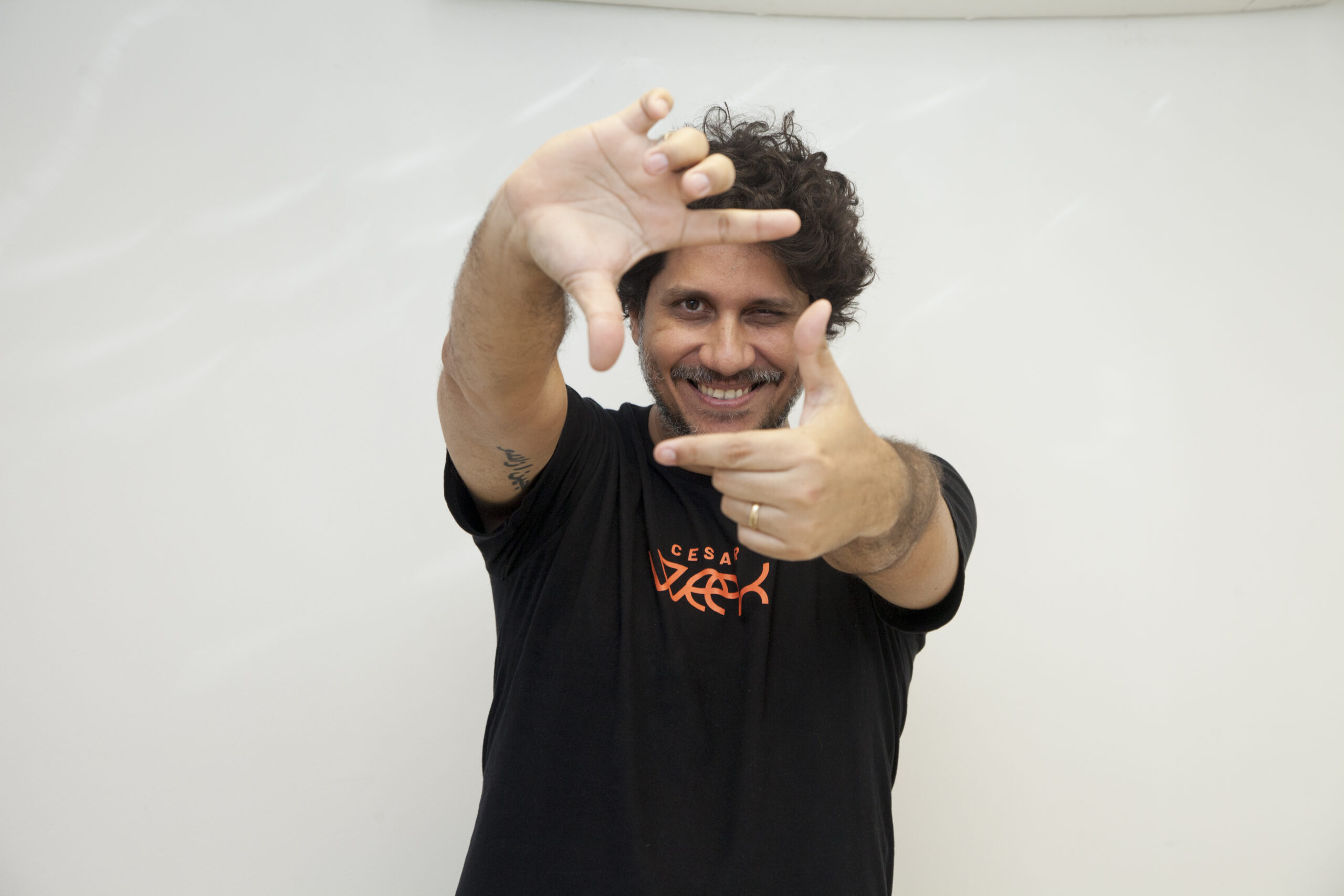 Homem sorridente com cabelos cacheados, usando camiseta preta, faz moldura com as mãos na frente do rosto contra fundo branco.
