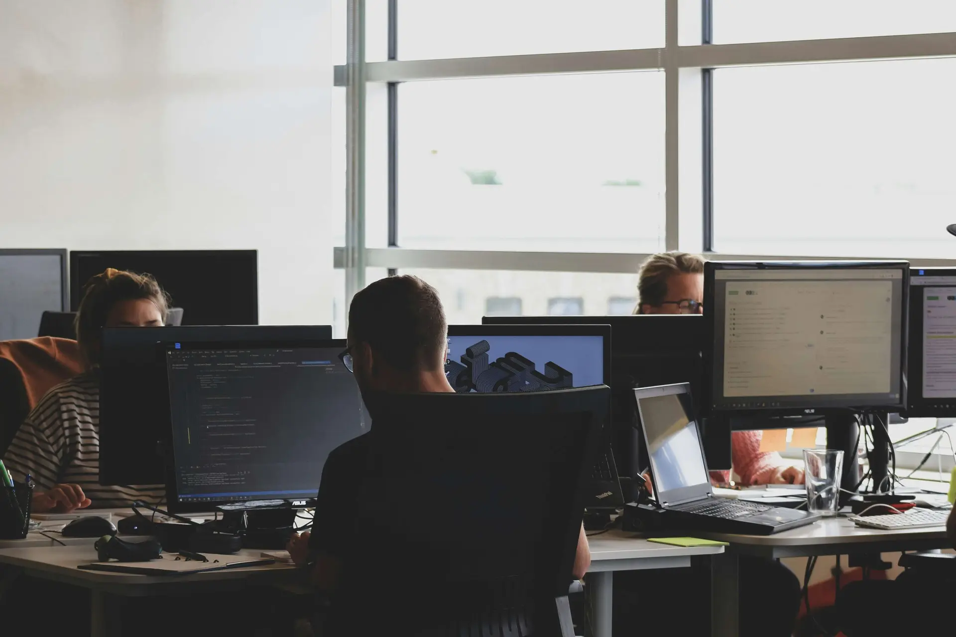 Três desenvolvedores trabalhando em computadores com linhas de código e gráficos em escritório iluminado com grandes janelas.