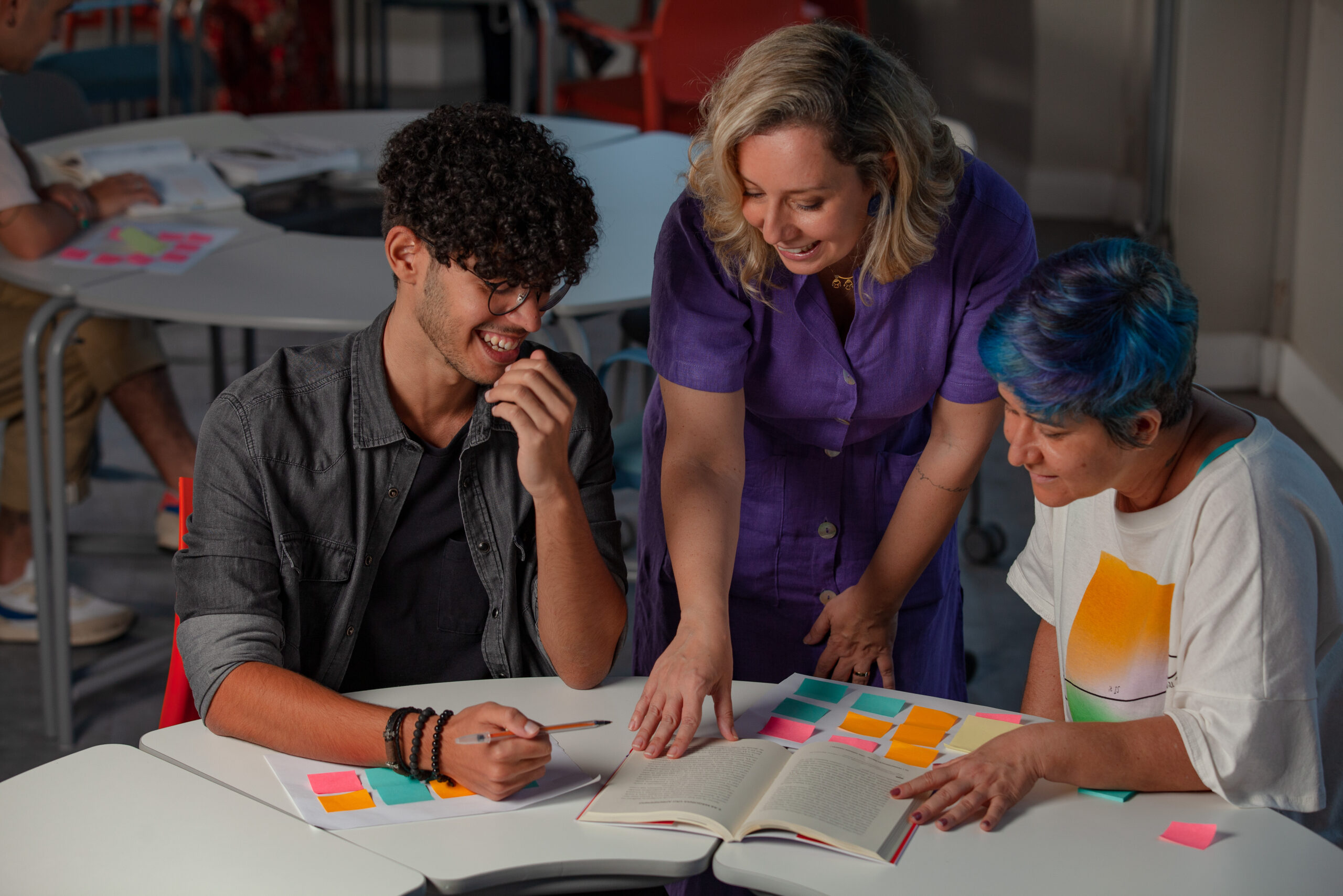 Três pessoas, uma professora e dois estudantes, sentados à mesa, revisando um livro e anotações coloridas.