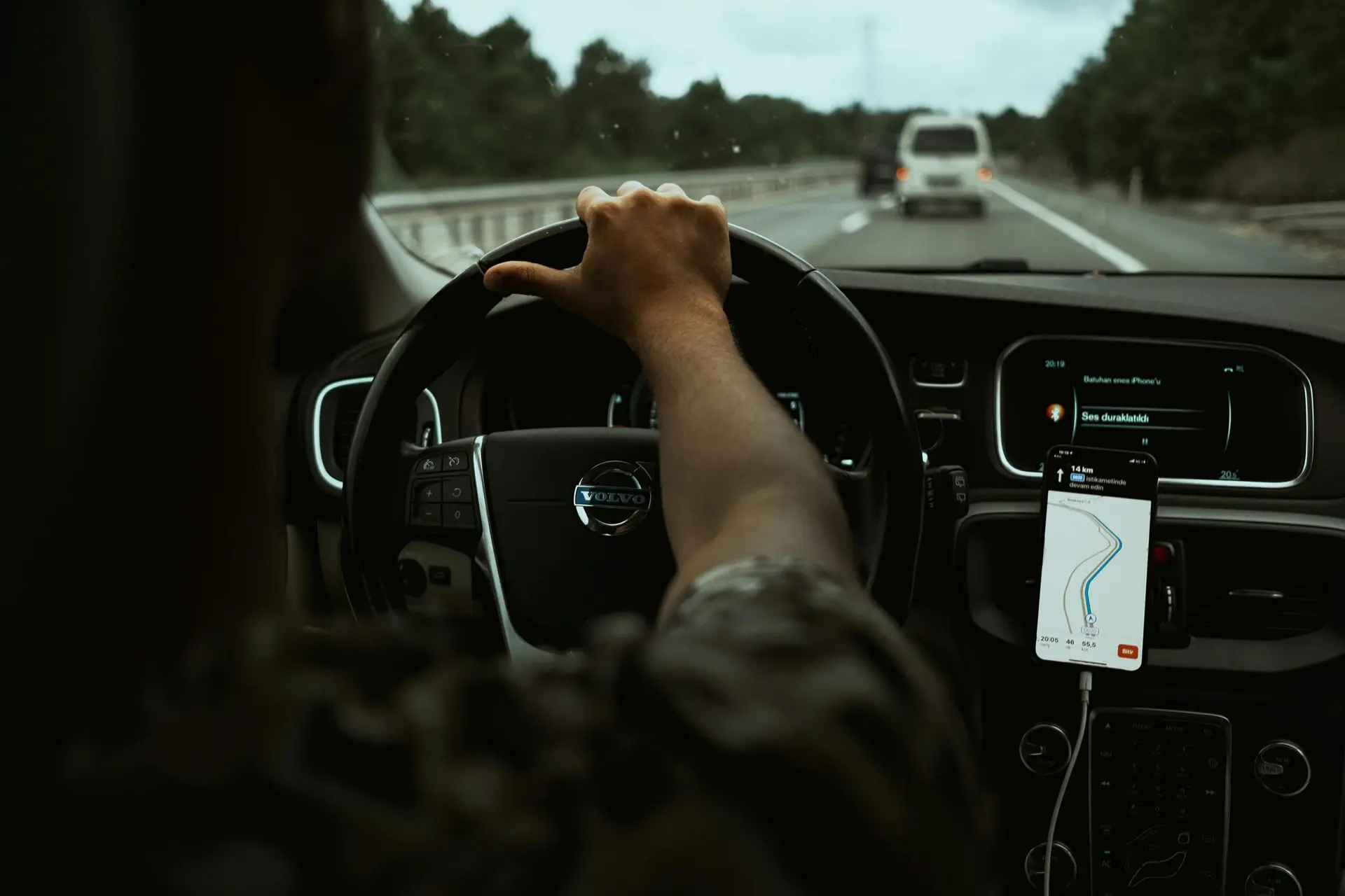 Visão interna de motorista conduzindo carro em rodovia, com smartphone exibindo mapa de navegação no painel central.