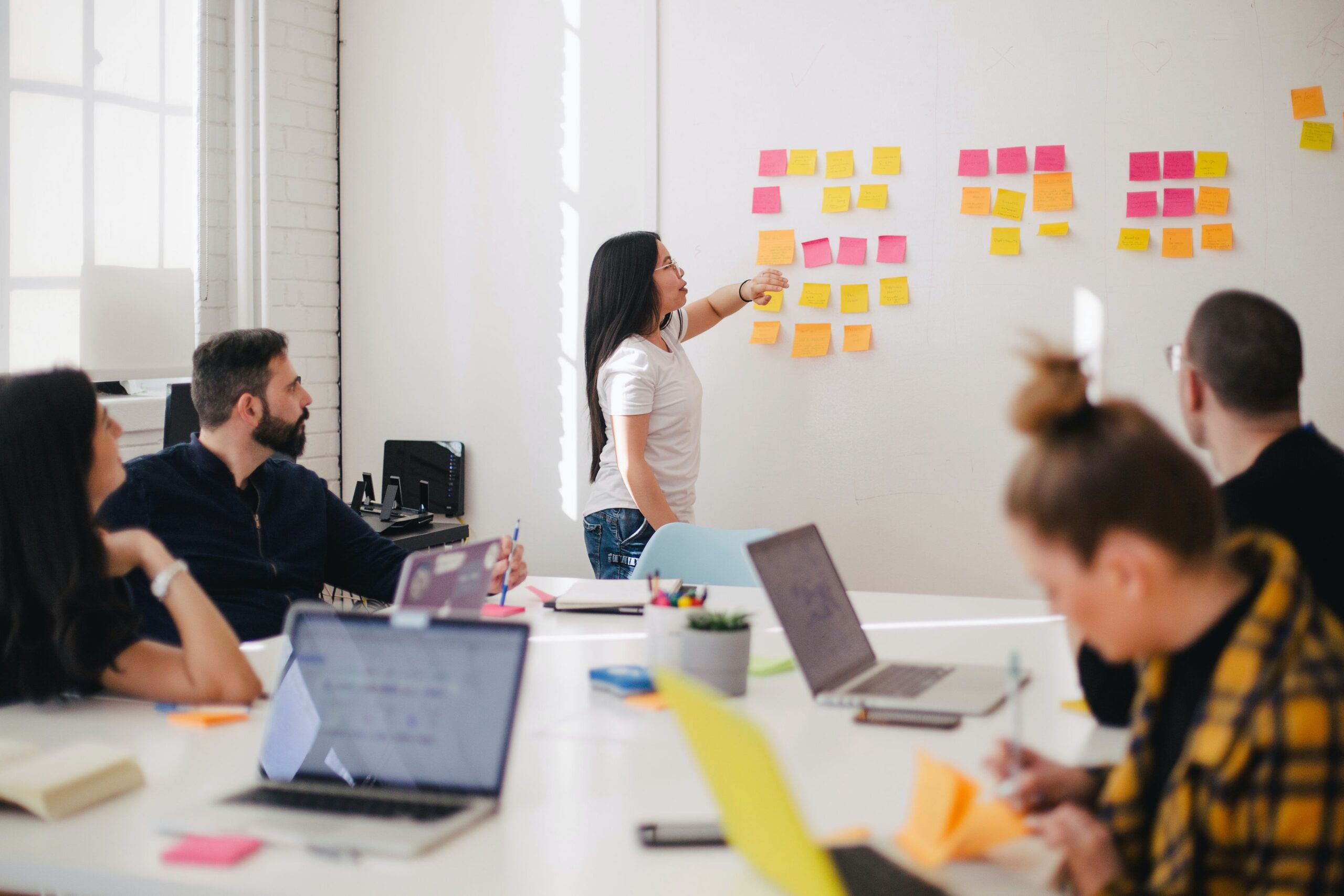 Uma mulher apresenta ideias em post-its coloridos a um grupo de colegas sentados em torno de uma mesa, com laptops abertos.