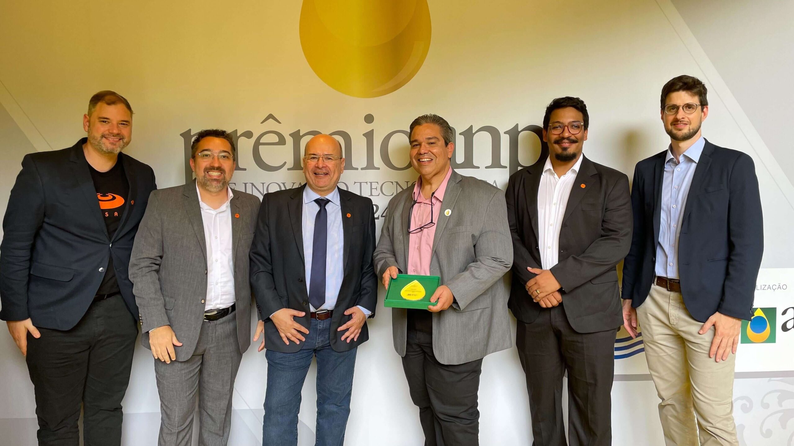 Cinco homens em ternos posam para uma foto em frente a um banner de premiação. Um deles segura um prêmio em forma de gota.