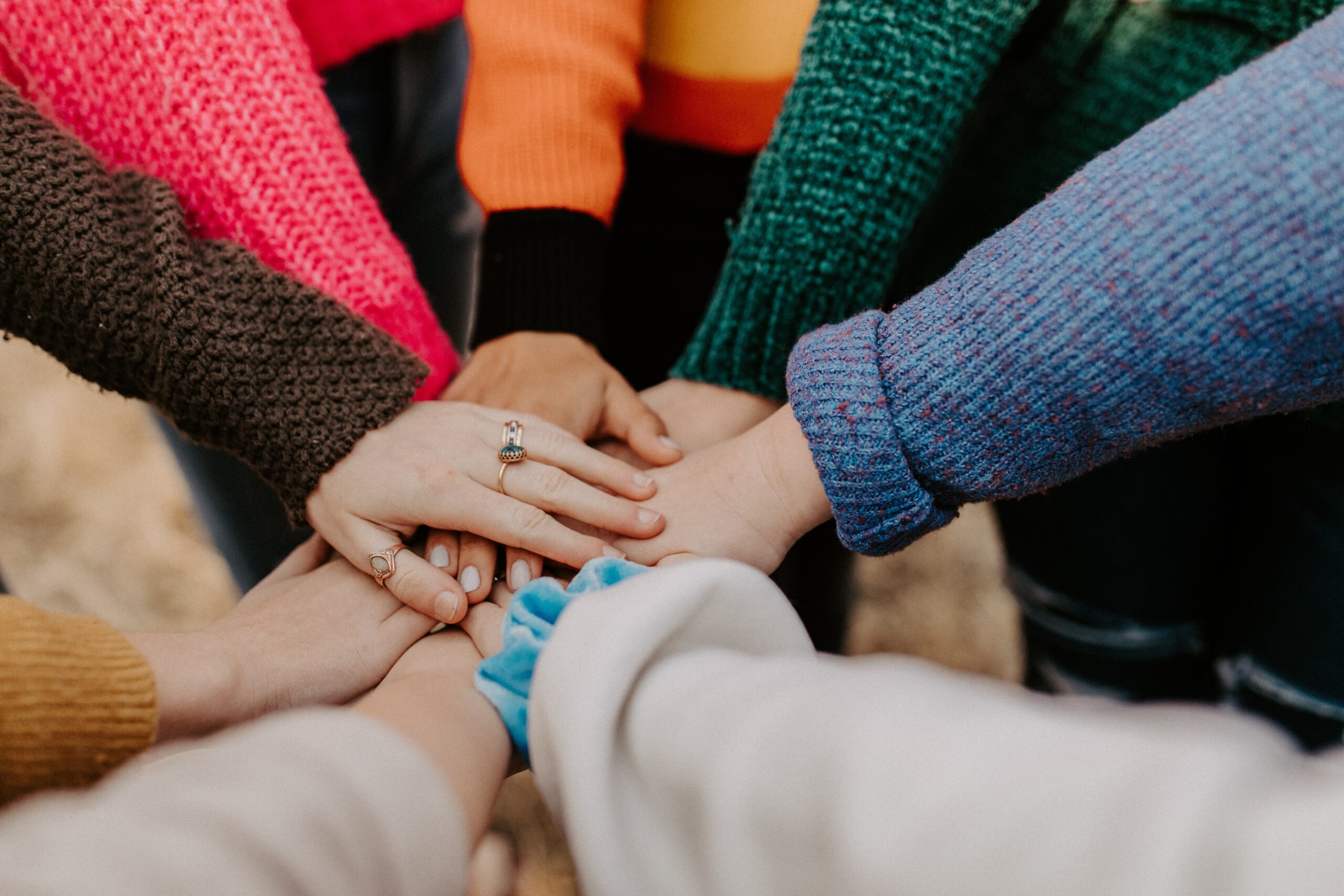 Mãos de diversas pessoas sobrepostas, mostrando unidade e trabalho em equipe. As pessoas vestem roupas de lã coloridas. Detalhe de anéis em algumas mãos.