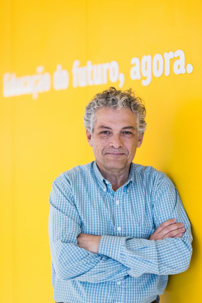 Homem de meia idade com camisa azul claro, braços cruzados, em frente a uma parede amarela com a frase "Criação do futuro, agora."