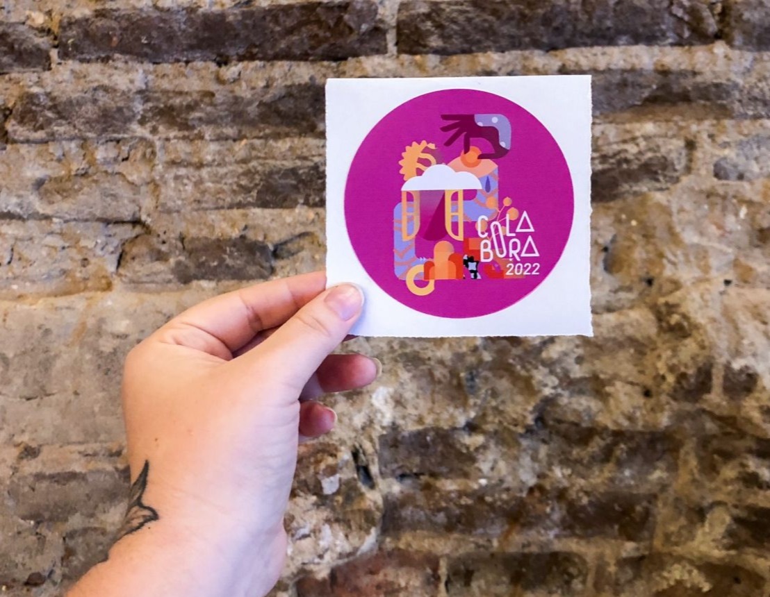 Uma mão segura um adesivo circular magenta com ilustrações abstratas e o texto "COLA BURA 2022". O fundo é uma parede de tijolos.
